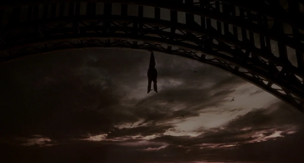 The Ladykillers (2004) movie still: wide — A dark, arched structural span looms over a cloudy night sky. A singular, pointed object hangs motionless…