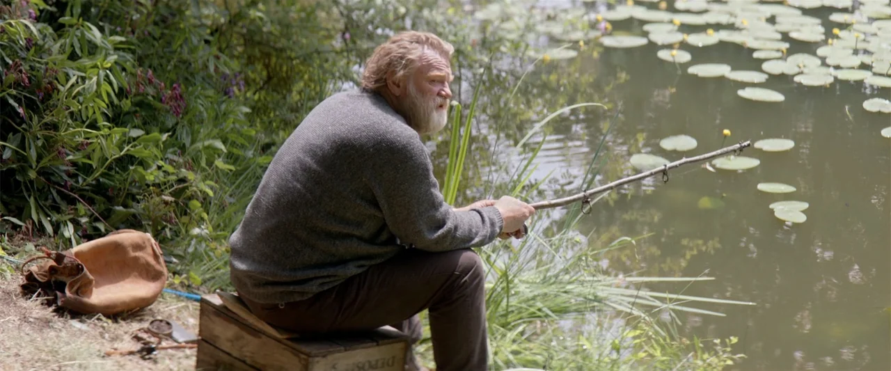 Hampstead (2017) movie still: medium — A quiet pond bank features a man sitting on a weathered wooden crate. The man holds a rudimentary stick…