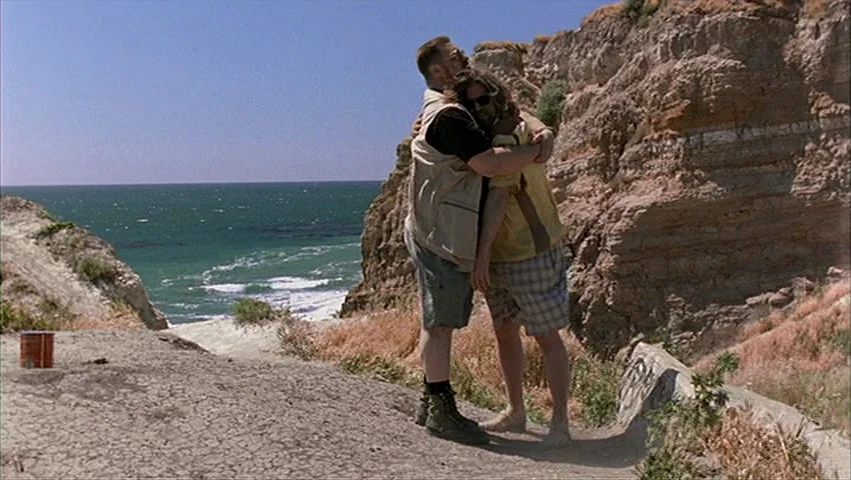 The Big Lebowski (1998) movie still: fullbody — A rugged cliffside overlooks the ocean with layered rock formations and sparse dry vegetation. Two…