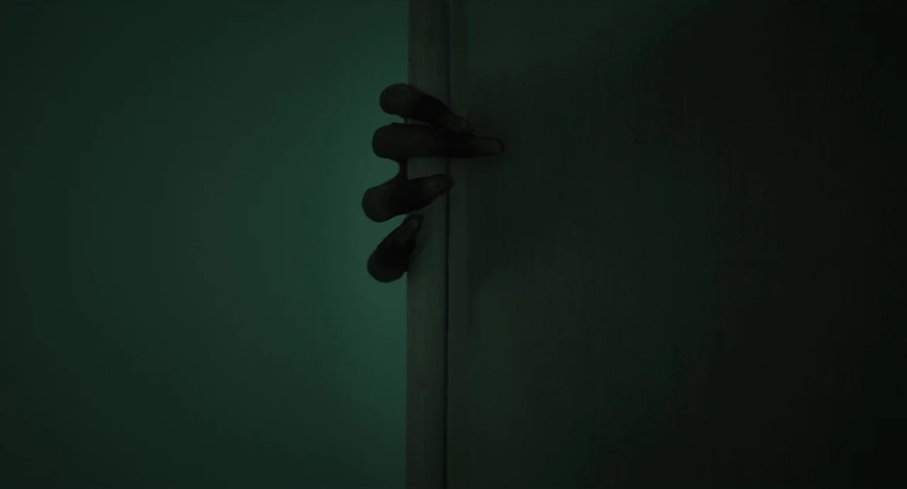May The Devil Take You (2018) movie still: closeup — A dark, plain interior wall is split by a vertical doorframe. A singular, dark-colored, clawed…