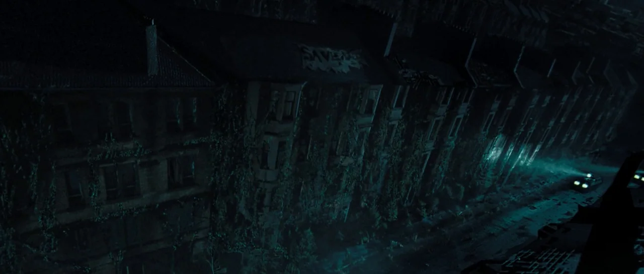 Doomsday (2008) movie still: establishing — A row of dark, ivy-covered tenement buildings lines a wet, narrow urban street at night. A lone vehicle…