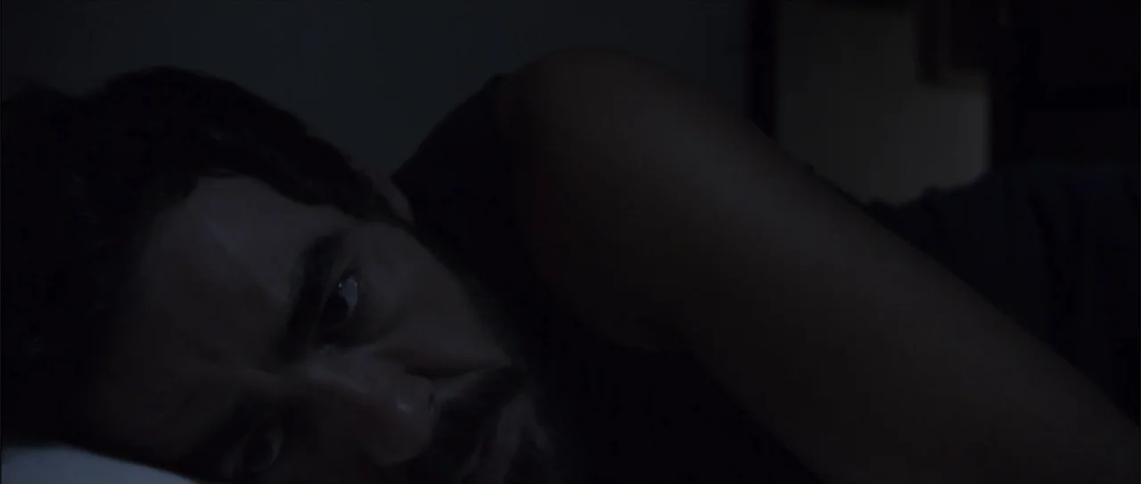 Otro verano (2013) movie still: closeup — A dark bedroom interior with a man lying on a white pillow. The man rests on the bed while looking toward…