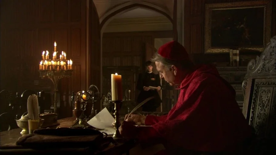 The Tudors (2007) movie still: medium — A dark, wood-paneled room is lit only by flickering candles on a desk. A cardinal writes at the desk while a…
