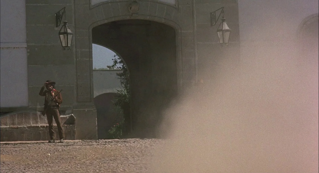 Bring Me The Head of Alfredo Garcia (1974) movie still: wide — A stone archway connects sections of an old courtyard with hanging lanterns. A man in…