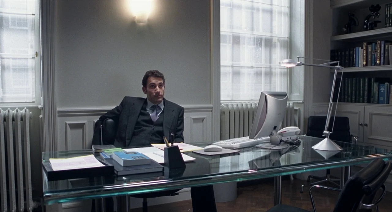 Closer (2004) movie still: medium — An office with a glass desk, grey walls, and windows with white curtains. A man sits behind the desk facing…