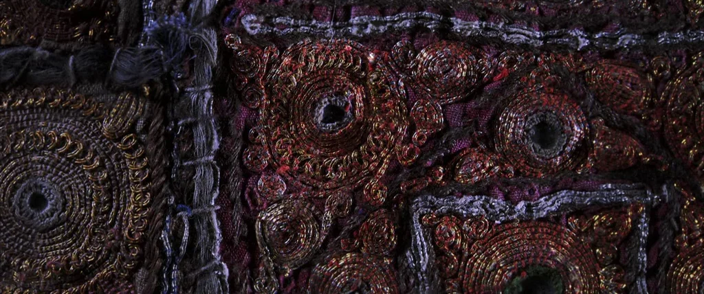 The Cell (2000) movie still: closeup — A close-up view of heavy, dark fabric adorned with intricate, circular gold-threaded embroidery patterns. The…