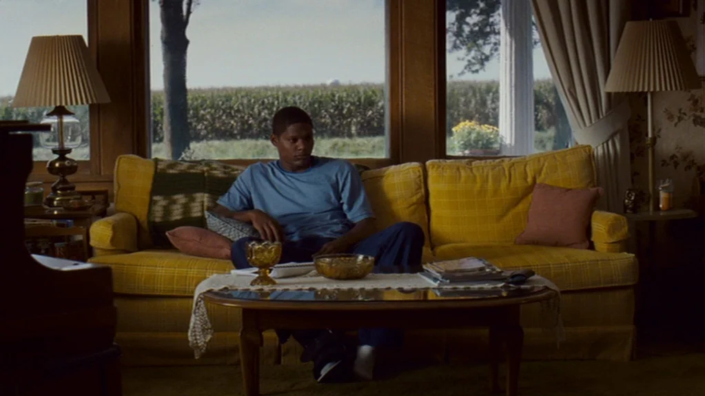 Sugar (2008) movie still: medium — A living room with a large yellow couch sits in front of a wide window. A young man sits in the middle of the…