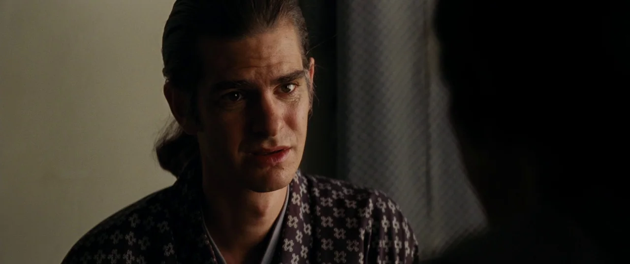 Silence (2016) movie still: closeup — A dimly lit room with dark wooden paneling and a visible patterned screen. A man with pulled-back dark hair…