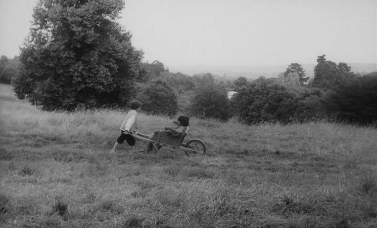 The Wild Child (1970) movie still: wide — A vast green meadow with tall grass under a pale, overcast sky. A child pushes another seated child in a…