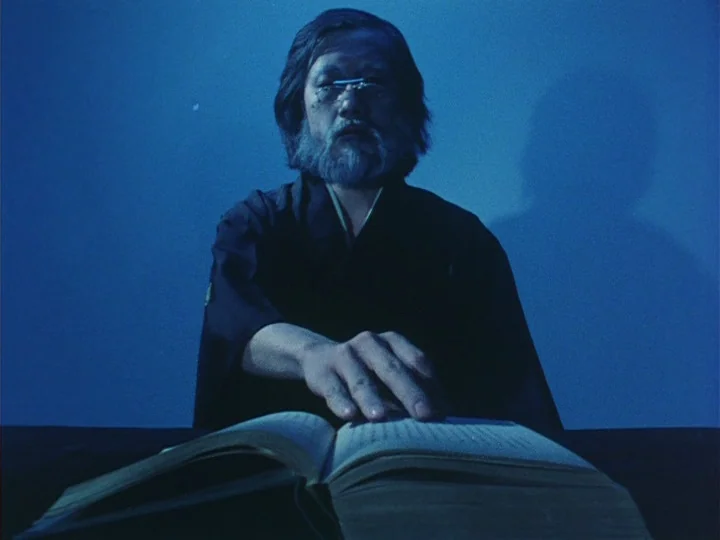 Trial (1975) movie still: medium — A man sits at a dark table against a plain, solid blue backdrop. He rests his right hand on the pages of an open…