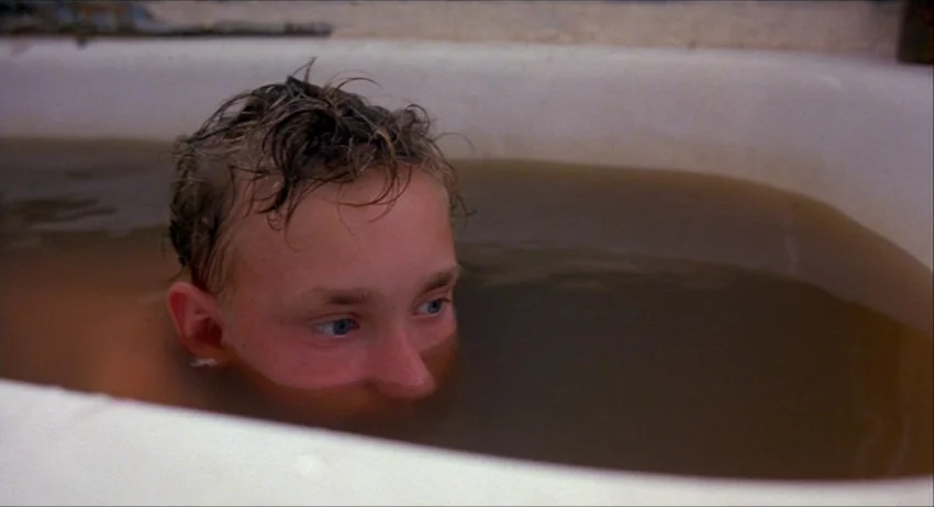 Gummo (1997) movie still: closeup — An off-white ceramic tub is filled with dark, opaque water. A young person with wet blonde hair rests in the…