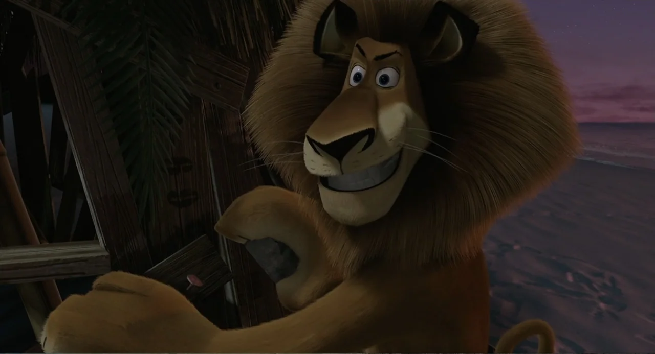 Madagascar (2005) movie still: medium — A makeshift wooden wall stands on a sandy beach during the late dusk hours. A male lion holds a rock while…