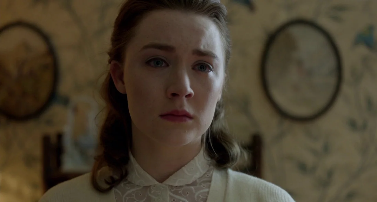Brooklyn (2015) movie still: closeup — A dimly lit room features patterned wallpaper with two blurred oval framed pictures. A woman with tear-filled…
