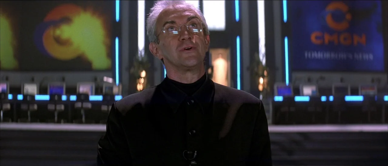 Tomorrow Never Dies (1997) movie still: medium — A high-tech media studio features large glowing orange logos on dark paneled walls. A man in a dark…