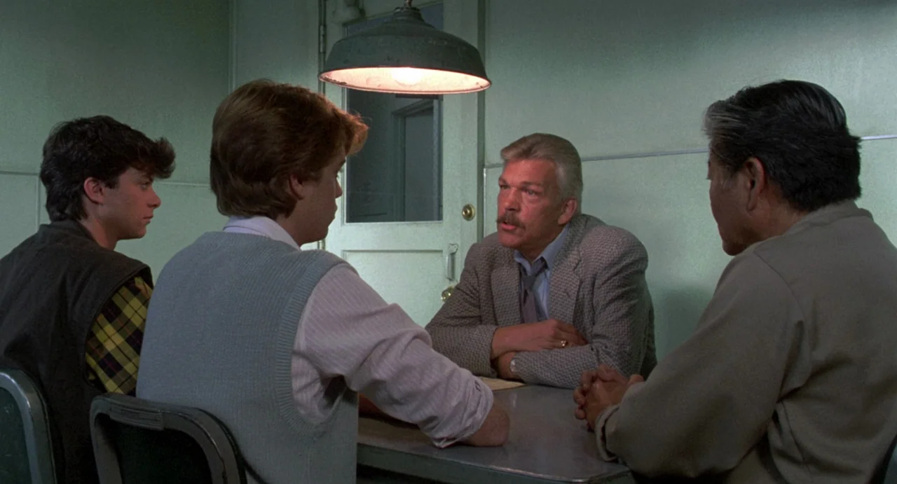 Night of the Creeps (1986) movie still: medium — A spartan room with light green walls contains a single metal table and door. Four men sit around…