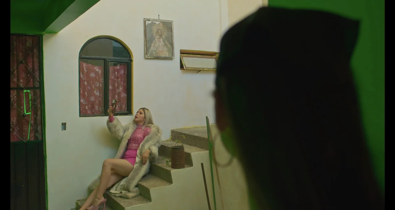 National Family (2023) movie still: medium — A woman in a faux fur coat poses for a photo on concrete steps. An out-of-focus individual is…