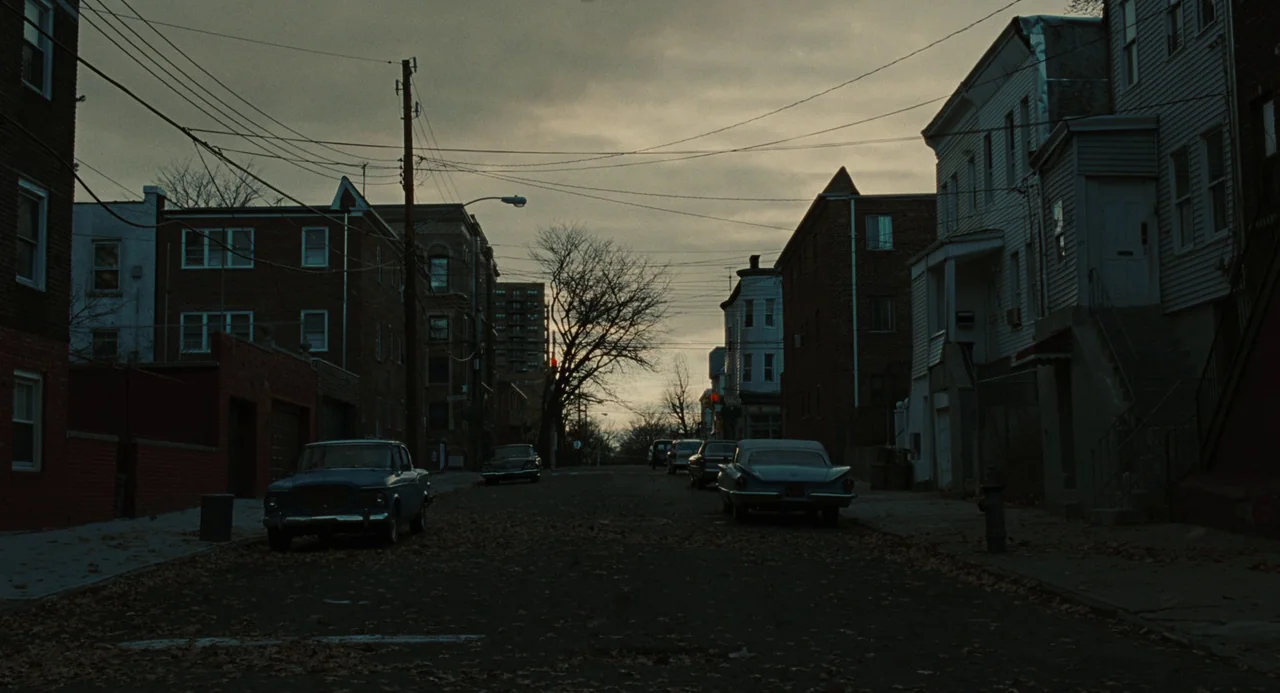 Doubt (2008) movie still: establishing — A quiet residential city street is lined with vintage cars and multi-story brick buildings. The pavement is…