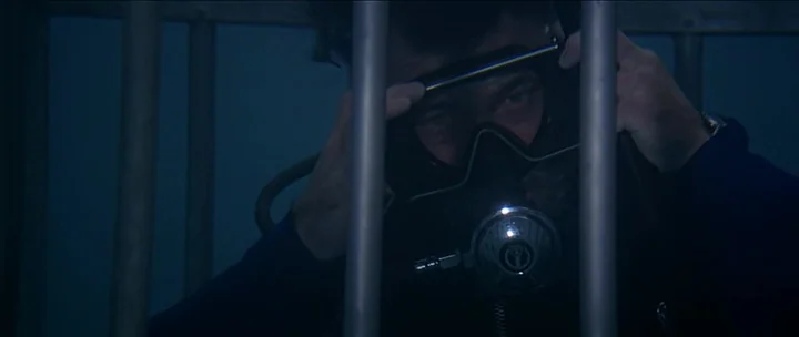 Jaws movie still: closeup — A person is inside a submerged metal cage with vertical bars. The individual adjusts a scuba mask and respirator while…