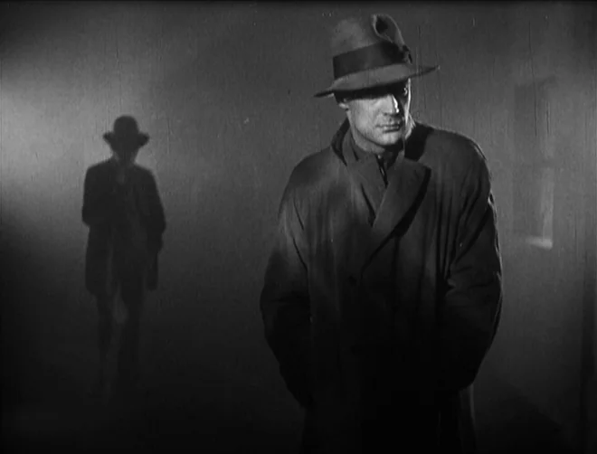 Pandora’s Box (1929) movie still: medium — A dark, dimly lit interior space features a foreground man in a classic trench coat. A distinct…