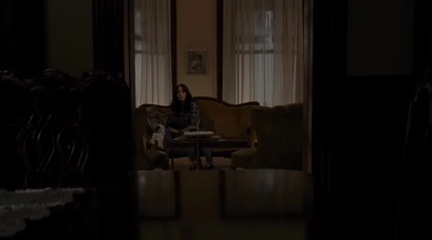 The House Of The Devil (2009) movie still: medium — A dark, dimly lit room with a woman sitting alone on a vintage sofa. Sunlight filters through…