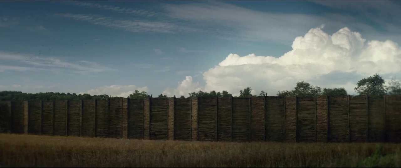 One & Two (2015) movie still: establishing — A tall woven stick fence spans across a dry golden field under blue skies. Large white cumulus clouds…