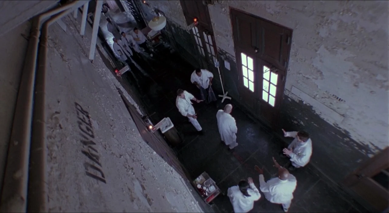 Twelve Monkeys (1995) movie still: wide — A long hospital hallway has peeling paint and a painted sign reading danger. Several people in white lab…