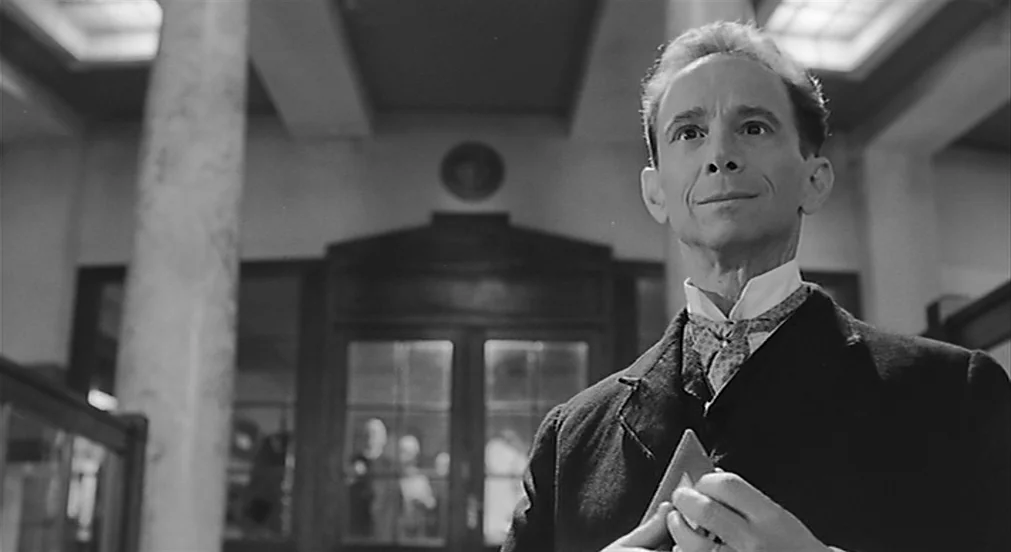 Kafka (1991) movie still: medium — A grand lobby with tall stone pillars and large glass pane doors. A middle-aged man in a formal suit stands…
