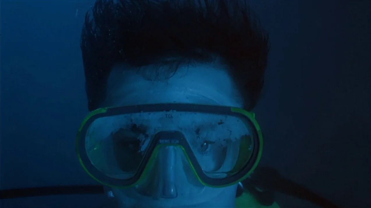 Dagon (2001) movie still: closeup — An underwater perspective shows a face obscured by a neon-trimmed diving mask. The surroundings are dark blue…