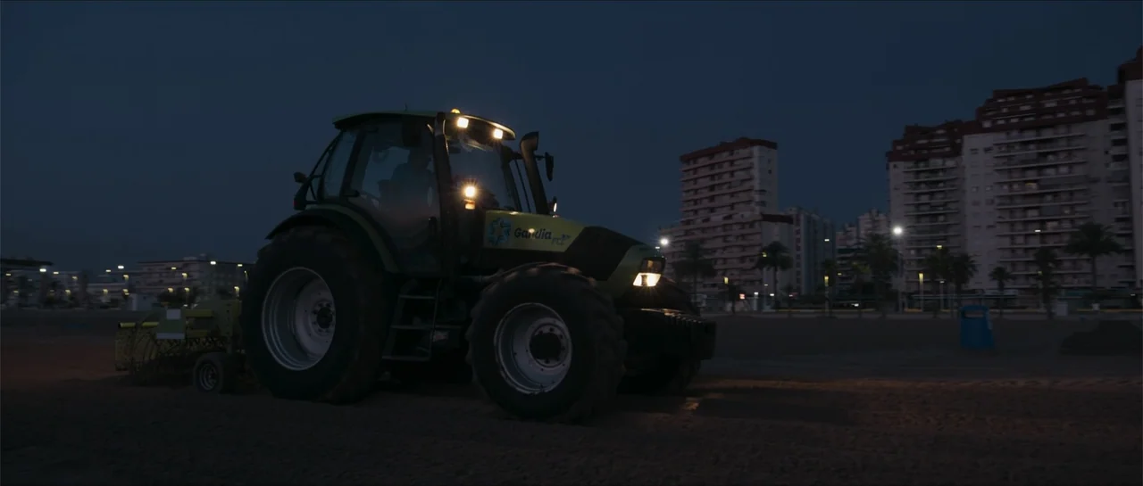 Historias lamentables (2021) movie still: medium — A green agricultural tractor is parked on a dark sandy beach at night. Residential apartment…