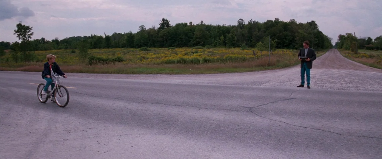 In The Mouth of Madness (1994) movie still: wide — A rural intersection where an asphalt road meets a gravel path under overcast skies. A young boy…