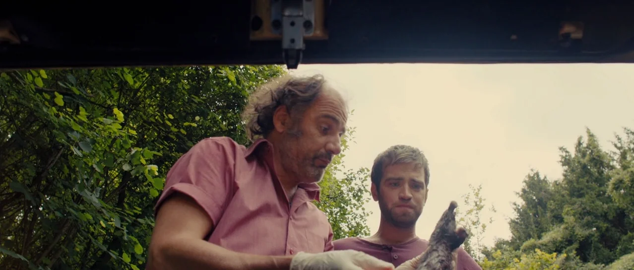 Road Games (2015) movie still: medium — Two men stand outdoors surrounded by lush green trees and foliage. One man inspects a deceased animal…