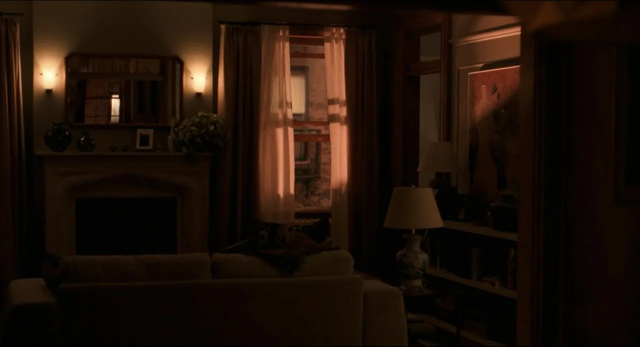 Karate Kid: Legends (2025) movie still: wide — A dimly lit living room with a fireplace and white sofa at night. Warm lamps illuminate the space…