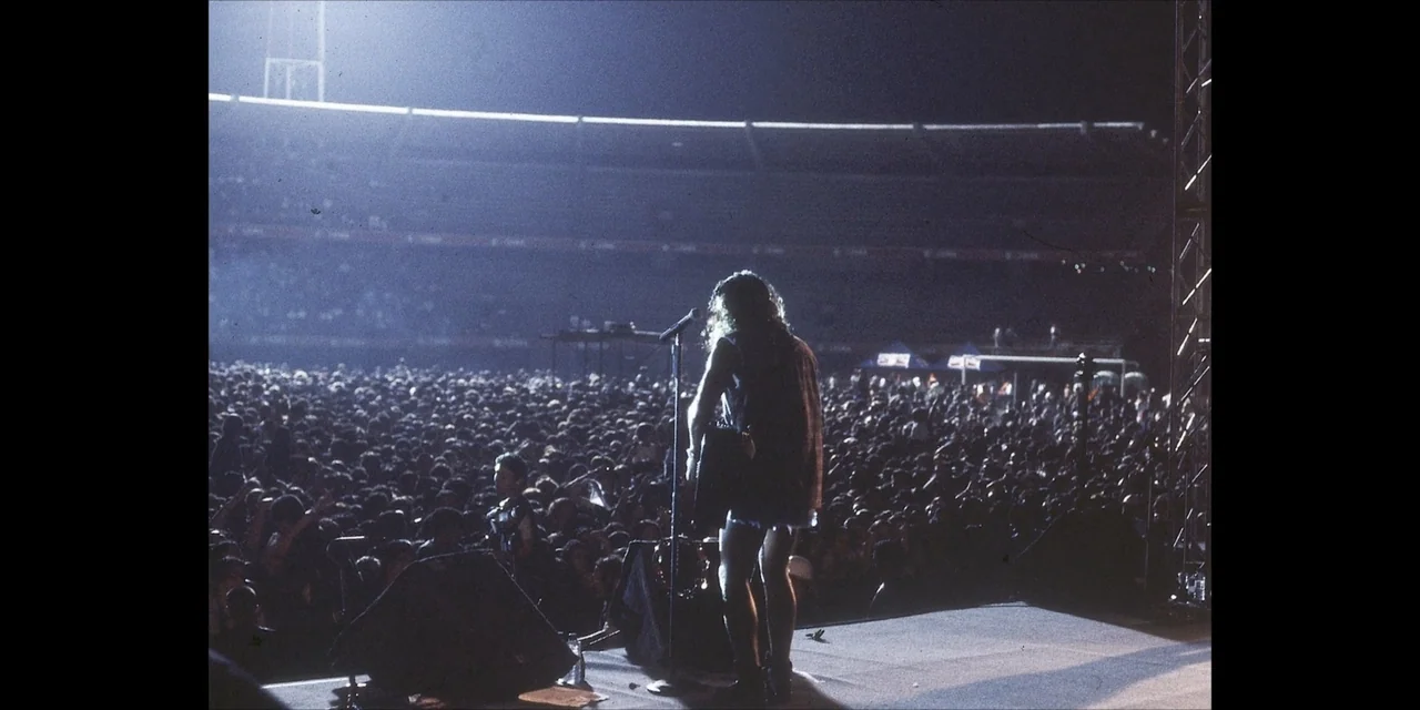 Leyenda Viva (2022) movie still: fullbody — A massive outdoor sports stadium at night packed with a dense, dark audience. A single long-haired…