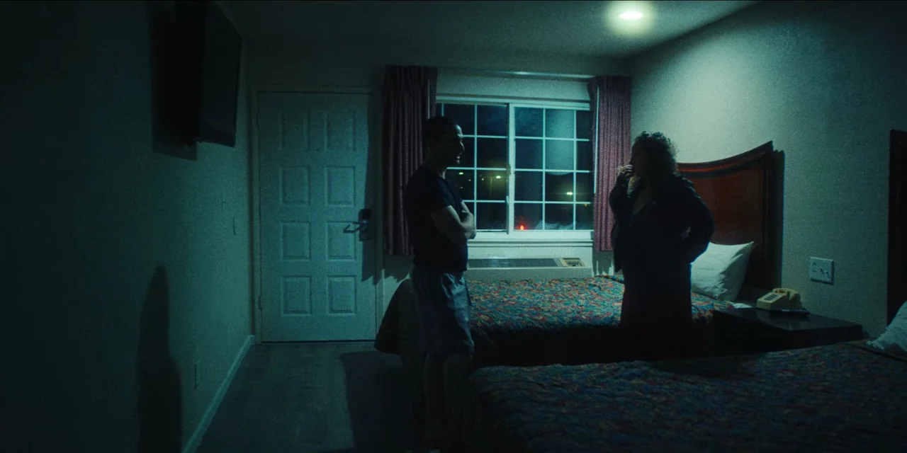 Spermworld (2024) movie still: wide — A dark, cramped motel room features two beds with patterned spreads and a single window. A man and a woman…