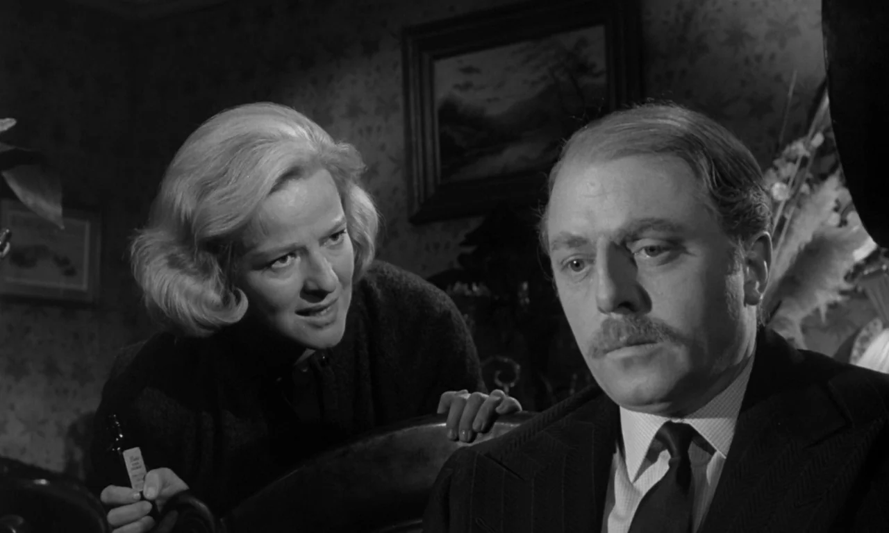 Seance on a Wet Afternoon (1964) movie still: medium — A dimly lit room with floral wallpaper and a framed landscape painting behind subjects. A…