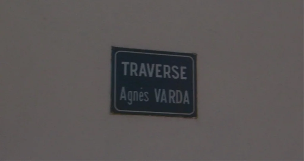The Beaches of Agnès (2008) movie still: closeup — A square metal street sign is mounted on a blank, off-white exterior wall. The sign displays the…