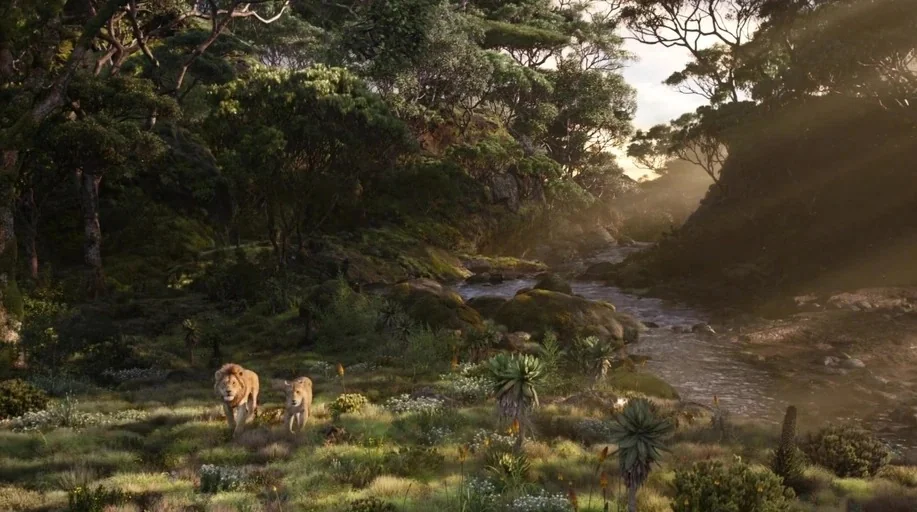 The Lion King (2019) movie still: wide — A lush river valley landscape features dense trees and rocky ground near flowing water. Two lions walk…