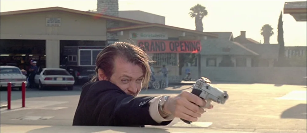 Reservoir Dogs (1992) movie still: medium — An outdoor gas station lot with a 'Grand Opening' banner hung on a building. A man in a dark suit leans…