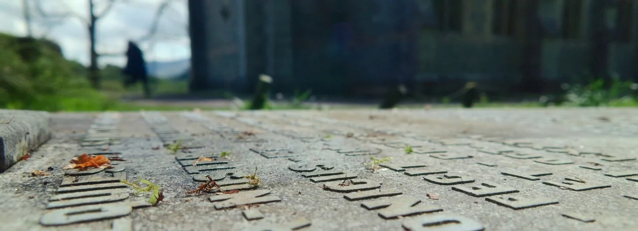 28 Years Later (2025) movie still: closeup — A stone grave marker features engraved lettering with moss growing inside the carved text. The…