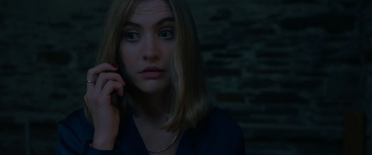 Oddity (2024) movie still: closeup — A dimly lit interior room features a dark stone or brick textured wall. A young woman with blonde hair listens…