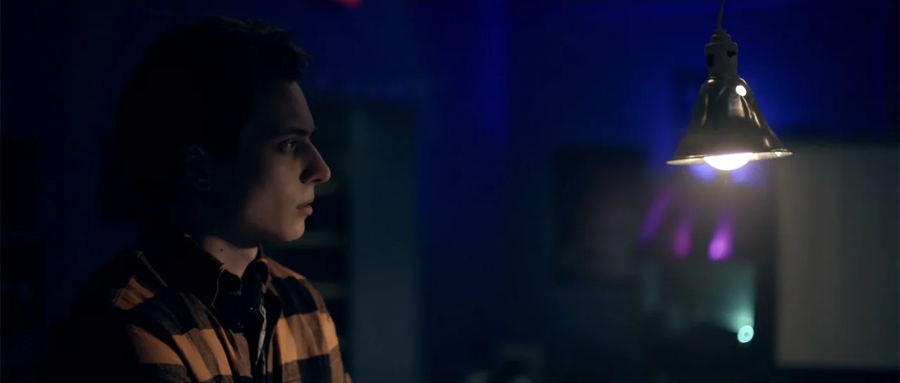 Elevator Game (2023) movie still: medium — A dimly lit room features a single hanging lamp as the main light source. A young man wearing a plaid…