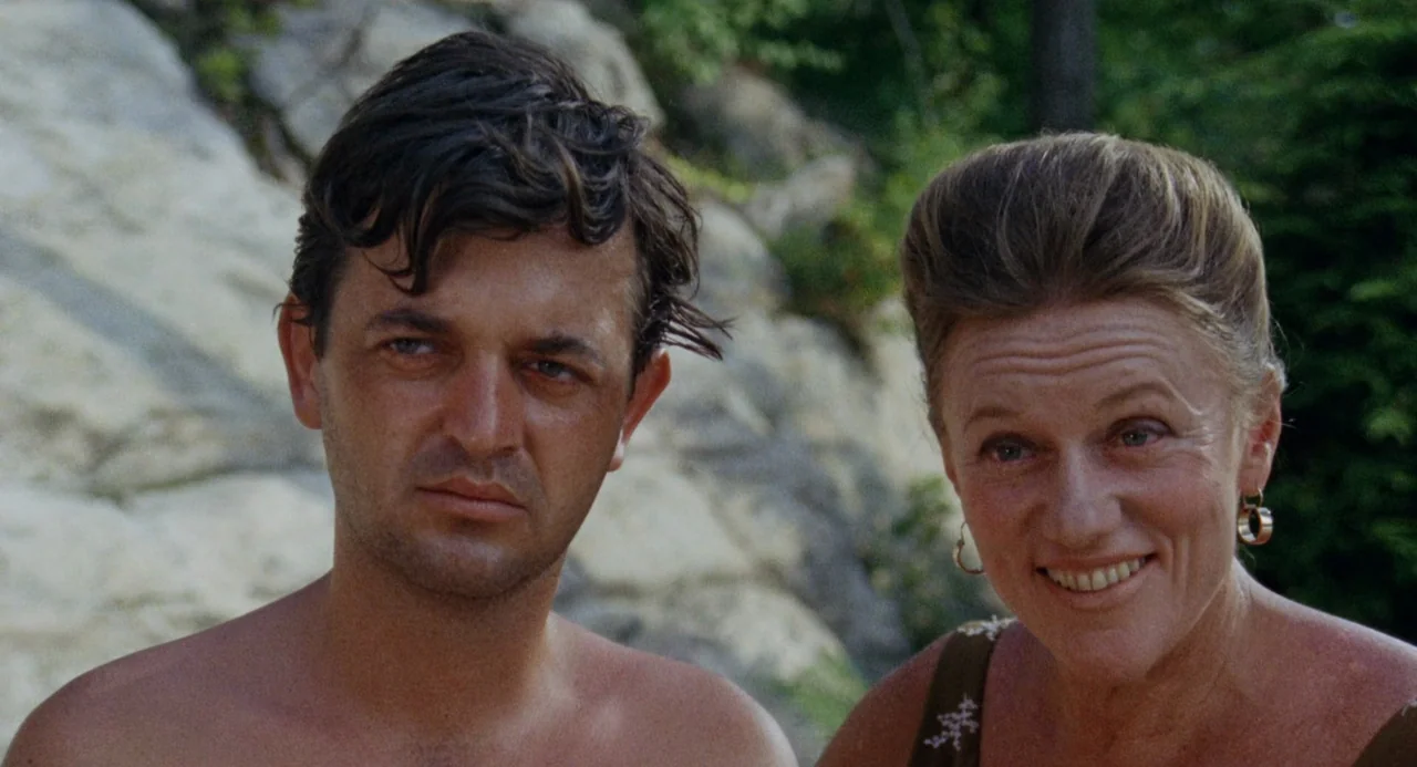 The Swimmer (1968) movie still: medium — A man and woman stand outdoors against a backdrop of pale, textured rock formations. The couple is…