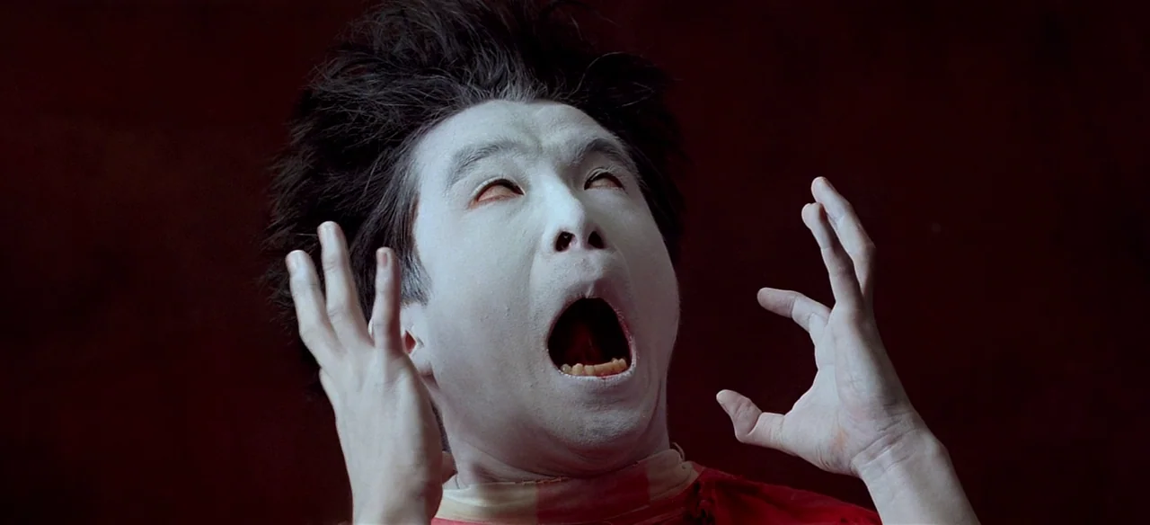 Baraka (1992) movie still: closeup — A person with white makeup stands against a dark, monochromatic red background. The subject holds their hands…