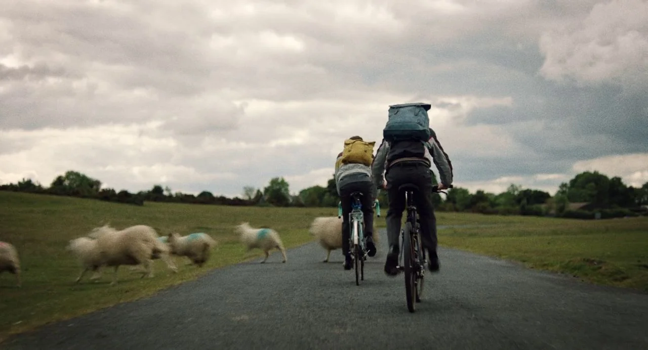 Dating Amber movie still: fullbody — A rural asphalt road cuts through a grassy pasture under a grey cloudy sky. Two teenagers ride bicycles away…