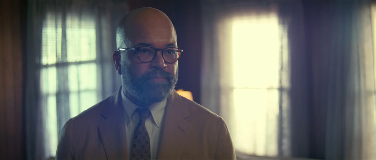 American Fiction (2023) movie still: medium — A man stands inside a dimly lit room with two bright windows behind him. He looks toward the right…