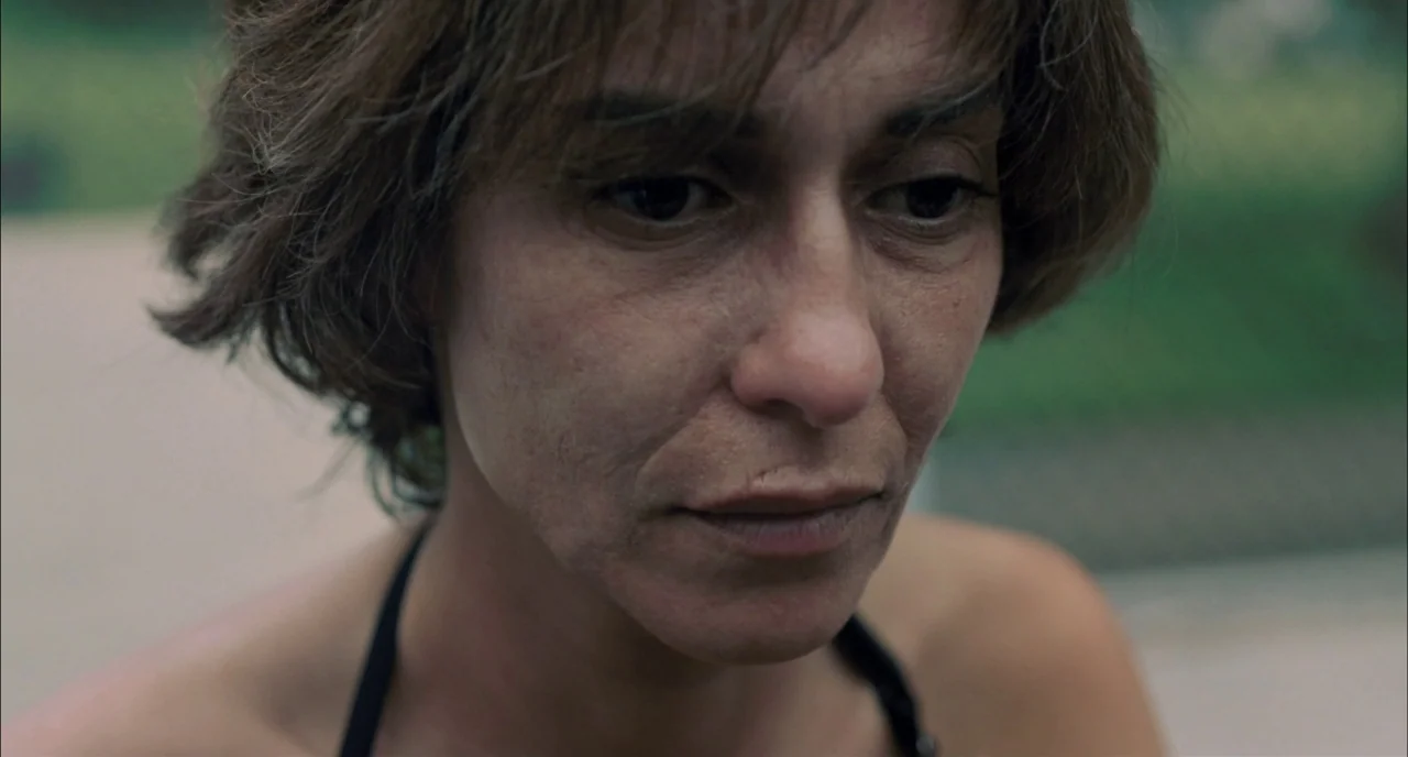 Incendies movie still: closeup — An outdoor park setting with blurred green foliage in the soft-focus background. A woman with short brown hair…