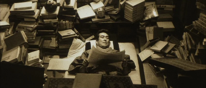 The Horrors of Malformed Men (1969) movie still: medium — A small room is filled with numerous precarious stacks of books on all sides. A man…