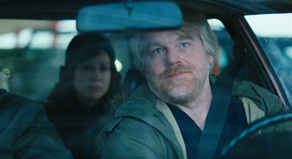 The Savages (2007) movie still: medium — The interior of a moving vehicle viewed from the front passenger seat perspective. A middle-aged man with a…