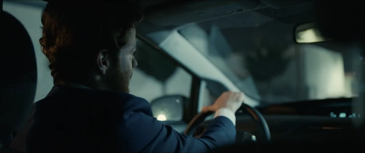 Plus One (2019) movie still: medium — An interior of a modern car at night looking over a driver's shoulder. A man in a suit drives while looking…
