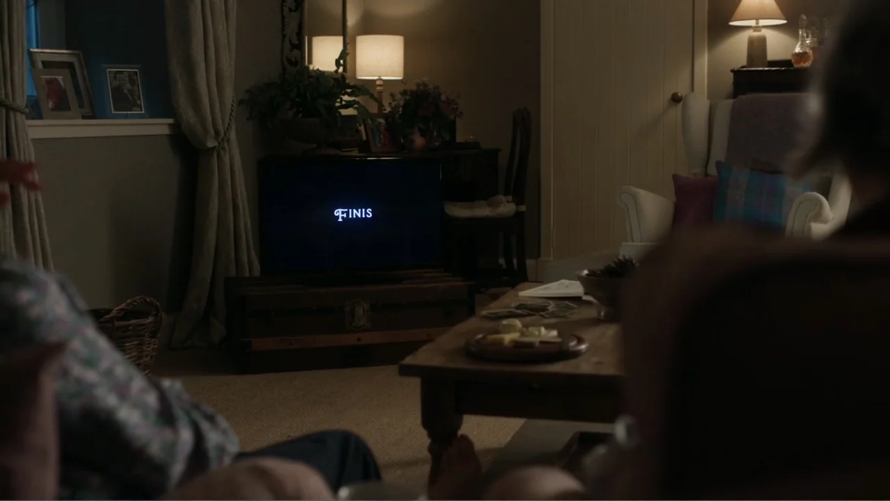 A Scottish Love Scheme (2024) movie still: medium — A traditional living room contains a television set displaying the word Finis on screen. Two…